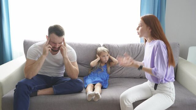 Caucasian child girl is suffering from quarrels between parents in family at home, redhead woman and bearded guy argue in presence of daughter.