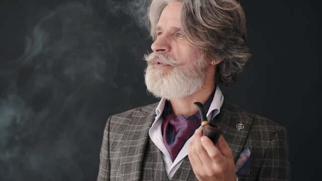Mature man smoking pipe and looking at it isolated on black background. Smoking habit. Fashionable mature man with pipe.