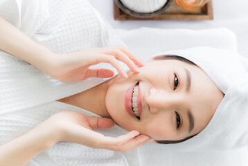 Fototapeta na wymiar Beautiful asian young woman touching her face by hands after clean fresh skin on face. Happy lady in white dress with lying down and enjoy of skin care treatment in spa salon. Skincare and spa concept