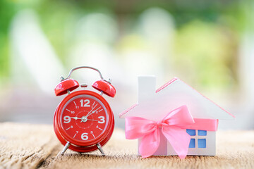 .Home model tied with pink ribbon and alarm clock on bokeh in the public park, The buy new house or real estate as gift to family or the one loved concept.