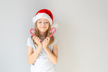 Portrait of a blonde caucasian smiling girl in a Santa hat holding two New Year's candies in her hands in front of her face on a grey background. Christmas and happines concept. Close up, copy space