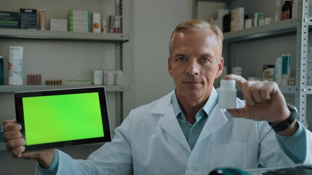 Portrait of a researcher Ph.D. with green screen Tablet PC and pills. Caucasian man, pharmacist. In white robe, smiles and holds a green screen digital Pad and dills. Place to insert your information.