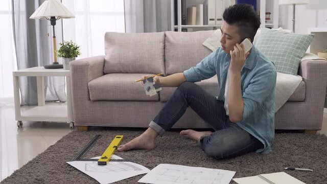 putting down housing model and taking up the blueprint, asian interior designer sitting on living room floor, reading drawing while communicating via phone.