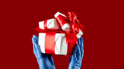 Christmas presents. Covid-19 protection. Greeting celebration. Safety delivery. Holidays 2021. Hands in medicine gloves holding gifts taped ruby ribbon isolated on red.