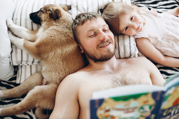 A man of European appearance reads a book for a night a girl of three years. The dog is sleeping on the bed. Father and daughter.