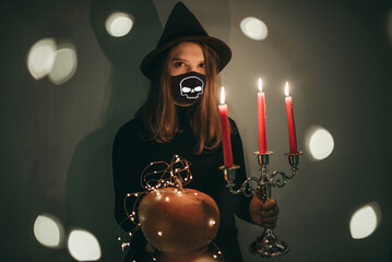 Young beautiful woman in a witch costume with candles, hat, magic, pumpkin in a medical black mask...