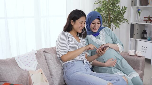 asian young woman and muslim expectant mom are having fun editing photos on the smartphone together in living room, laughing while looking and touching the screen.