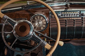 Interior close up of luxury, classic, vintage car with steering wheel, ivory gear shifter, chrome circular speedometer, radio, and wooden dashboard