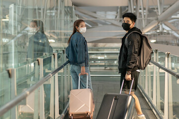 Two Asian traveller couple in casual clothes wearing face mask to prevent coronavirus walking with luggage in airport walkway. new normal and keep social distancing lifestyle. safety travel concept