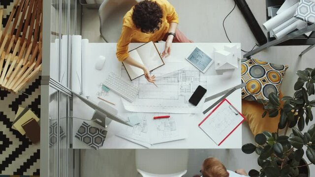 Top Down Shot Of Female Architect Using Digital Tablet At Office Desk, Then Greeting Male Colleague With Fist Bump And Discussing Floor Plan With Him