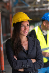 Smiling young woman construction manager or engineer posing with crossed arms and looking away. On background, out of focused construction worker