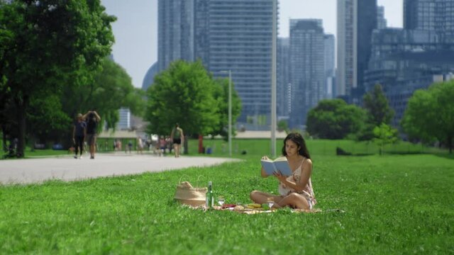 Curious young brunette drinking wine and reading a blue book at a busy urban green park on a warm summer day