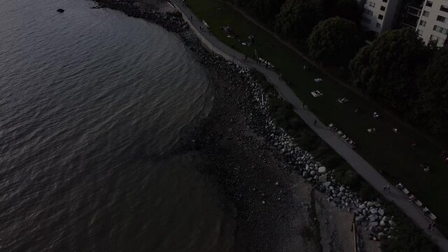 evening aerial over beach park panaramic pan out people alking on boardwalk sea wall path while couples and family seated on blankets towels with respectful social physical distancing West coast
