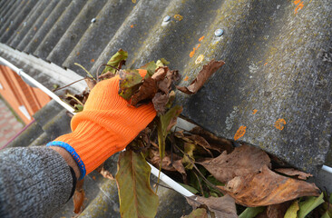 A man in gloves is cleaning a blocked rain gutter attached to the asbestos roof by removing fallen...