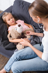 Selective focus of woman in stethoscope sitting near daughter with teddy bear on sofa