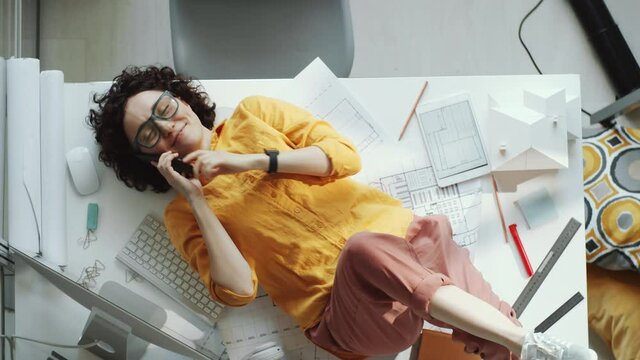 Top down shot of young positive female architect lying on blueprints on office desk, smiling and laughing while chatting on mobile phone during workday