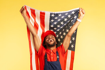 Extremely happy satisfied craftsman in uniform holding flag of united states of America, celebrating labor day, independence. Indoor studio shot isolated on yellow background