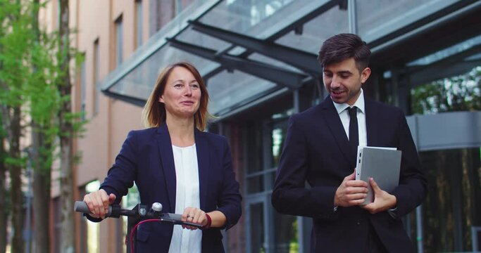 Portrait of Caucasian businessman and businesswoman walking on street. Adult female and male collegue with kick e-scooter and laptop talking and laughing. Technology, partnership, occupation.