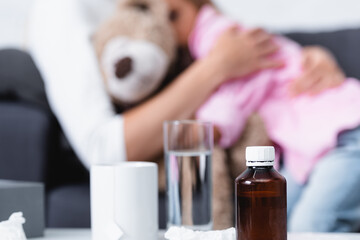 Selective focus of bottle of syrup, cup and glass of water near mother embracing kid