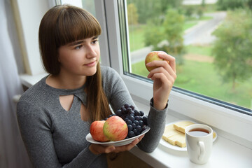 A young girl with a plate of fruit in her hand and a Cup of coffee looks out the window.