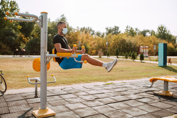 Fototapeta premium Side view of fit young male caucasian athlete with face mask trains on arm and leg equipment in an outdoor gym. Muscular athlete outdoors. COVID - 19 coronavirus protection