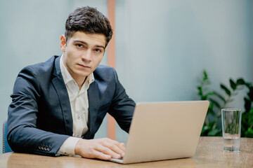 A young brunette man of European appearance commented on the message sits at a desk in front of a laptop, looks at the camera and thinks about a new project.