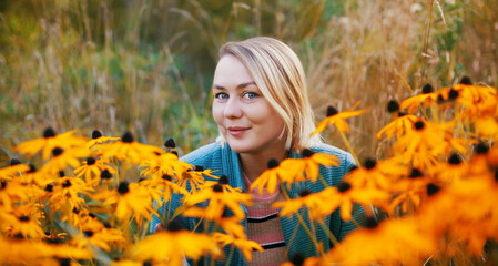 Smiling young woman sitting in the flowers. Blonde female autumn outdoor portrait with blooming flower meadow