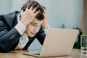 holding his head strongly worried male businessman of European appearance in a stylish black suit looking at a laptop monitor.