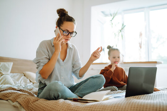 Young beautiful businesswoman talking on mobile phone and working on a laptop. Busy young woman with daughter in home office. Stay at home. Covid-19.