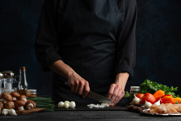 The chef in black uniform cuts with knife onion on black board for adds to the meal. Dark blue background. Preparing food concept.