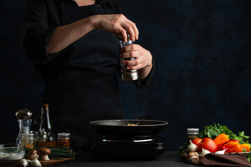 The chef adds black pepper to the dish into frying pan on professional kitchen. Dark blue background. Preparing restaurant meal. Food concept.