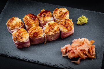 Served warm sushi rolled in bacon with ginger and wasabi on a black stone plate.