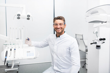 Confident male dentist sitting in a modern clinic near his dental chair. Dental hygienist wearing protective glasses smiling at the camera