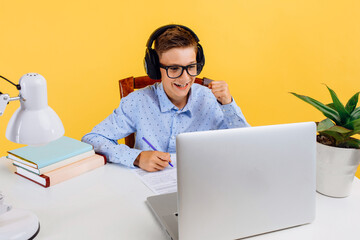 schoolboy guy in headphones, doing homework using a laptop computer, learning through an online...