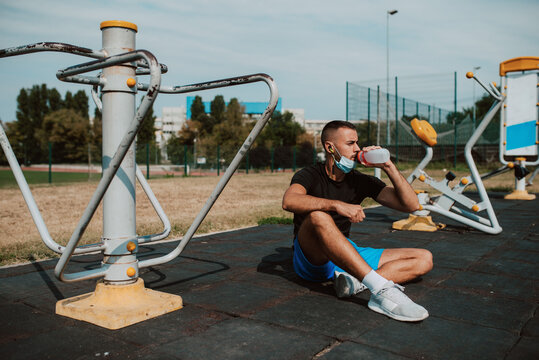 A fit muscular male caucasian athlete with a face mask sits next to a equipment in an outdoor gym and drinks water from a bottle. Coronavirus protection COVID - 19