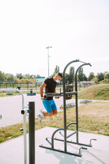 Fototapeta na wymiar Fit young male caucasian athlete with face mask trains on arm and leg equipment in an outdoor gym. Muscular athlete outdoors. COVID - 19 coronavirus protection