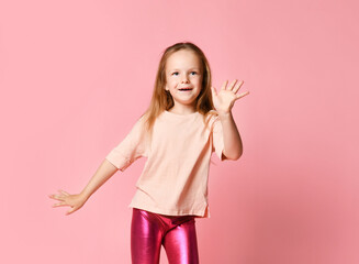 Cute blonde kid in t-shirt and leggings. She smiling and dancing or saying hi showing her palm, posing on pink studio background. Close up