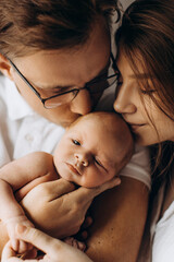 Happy parents with cute newborn baby girl, handsome dad hold little daughter in arms, beautiful wife and loving husband gently kiss their child, young family concept