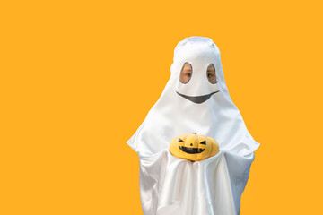 little boy looking out from ghost costume with smile holding orange pumpkin  with black painted face on  orange background. Halloween celebration concept. Copy space for text.