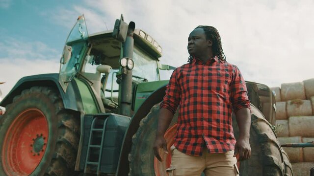 African man, farmer standing in front of the green tractor and crossing his hands. Hay rools stack in the background. High quality 4k footage
