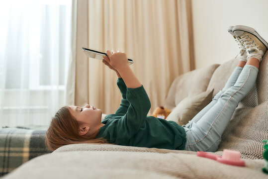 A small cute girl playing games on a tablet lying on a sofa with her feet up and gumshoes on