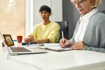 Cropped shot of aged woman, senior intern making notes while listening to her young colleague,...