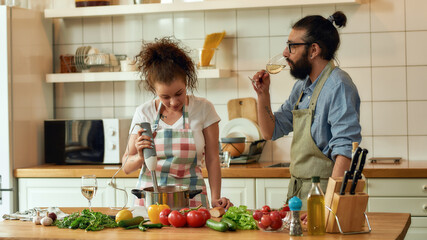 Italian man, chef cook drinking white wine while woman using hand blender. Young couple preparing a meal together in the kitchen. Cooking at home, Italian cuisine