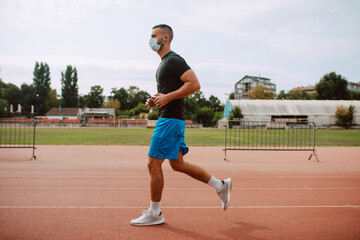 Fototapeta premium A shot of a young male caucasian athlete with face mask training on a racetrack. The sprinter runs on athletic all-weather running track. COVID - 19 coronavirus protection