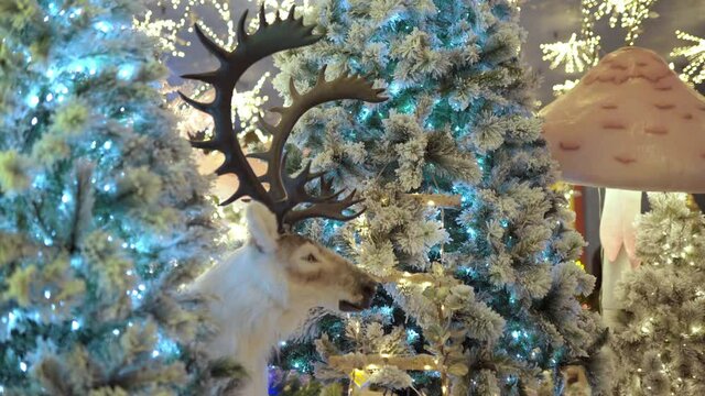 Majestic arctic white reindeer with amazing horns among the glittering christmas trees, christmas ornaments decorated with colorful led garlands. Christmas decorations on illuminated night street or