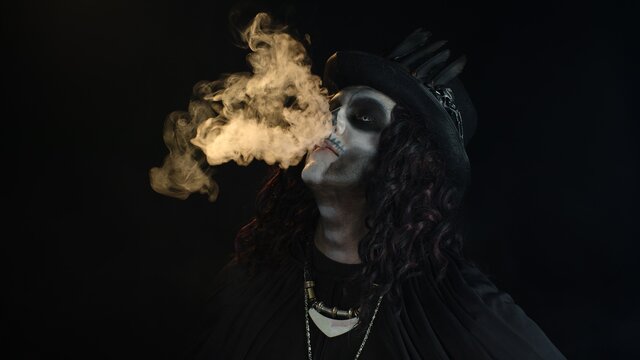 Sinister man with professional skull makeup exhaling cigarette smoke from his mouth and nose