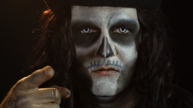 Creepy long hair man with skeleton makeup making faces and pointing at camera, trying to scare