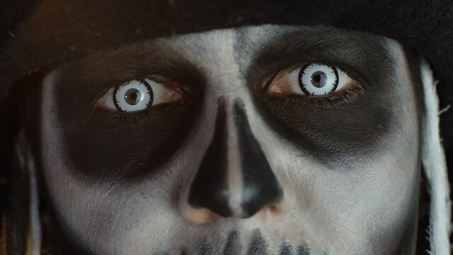 Close-up shot of face in skeleton Halloween makeup opening eyes with white pupil, trying to scare