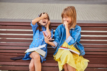 Two multinational 9-year-old girls are sitting on a bench with phones, they communicate with each...
