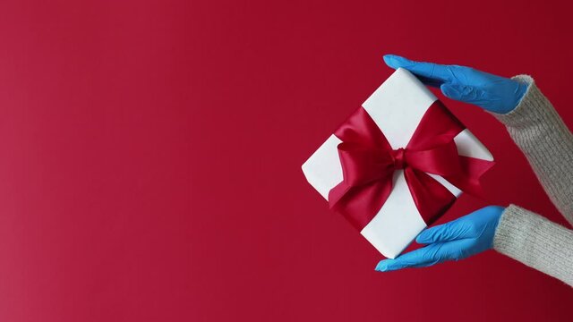 Winter holidays gift. Pandemic Christmas 2021. Coronavirus quarantine special day surprise. Hands in blue gloves holding present in white box with ribbon bow isolated on red copy space background.